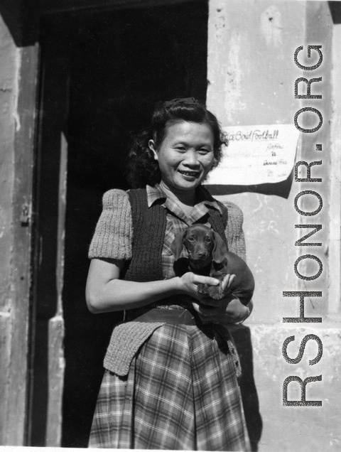 Alice Chong outside the HQ building at Kunming in 1944, holding the Dachshund puppy that her roommate Eloise Witwer gave to General Chennault.  Chennault named the dog “Little Joe” and then changed its name to “Joe-Dog” after it grew up.  The name, of course, was a way of getting back at his nemesis, General Joseph Stilwell because, as Chennault would often say, “Both were low-down, sons of bitches”. . .  Note the notice for the upcoming “Rice Bowl” football game pinned to the door frame too.  This is likel