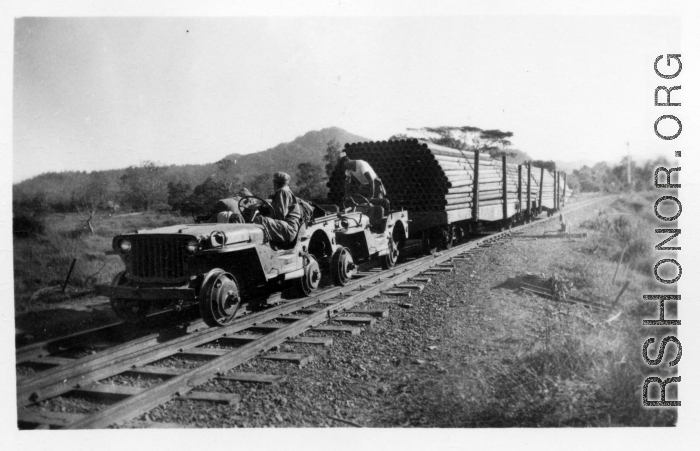 Two jeeps, fitted with steel wheels that fit the rail tracks, pull cars loaded with oil pipes. In Burma.  During WWII.  797th Engineer Forestry Company.