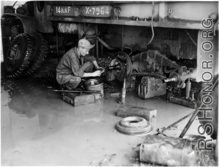 An American GI repairs a transport truck of the 14th Air Force in China, working in rough conditions on flooded ground.