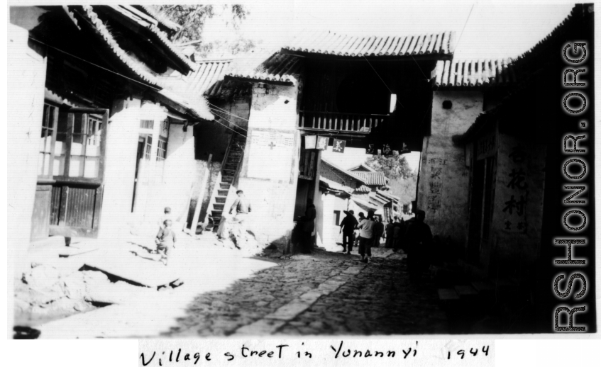 Village street in Yunnanyi, China, during WWII, with gate having a sign for Shanghai Man Hairstyling (上海男美发室), and the Almond Flower Tea Room (杏花茶室）in the shadow on the right. 1944.