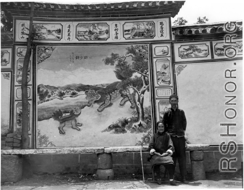 An elderly woman and man pose before a traditional decorative painted wall in SW China during WWII.
