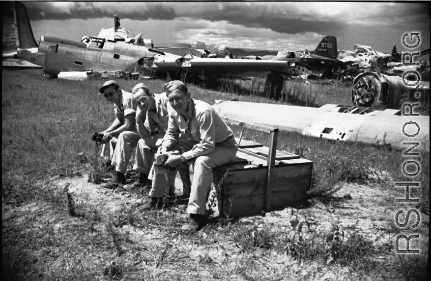 Three American GIs sitting on a discarded latrine box in front of wrecked American warplanes (including a B-24) in a boneyard at the base. An aircraft tail #349752 is in the background. Note men walking on wing of aircraft in background, undoubtedly engaged in salvage on the wing. In Yunnan, China, during WWII.