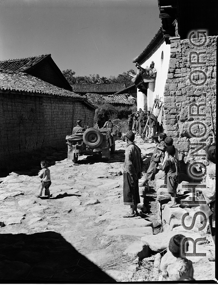 GI drives jeep in village lane among adobe brick houses in Yunnan, during WWII.