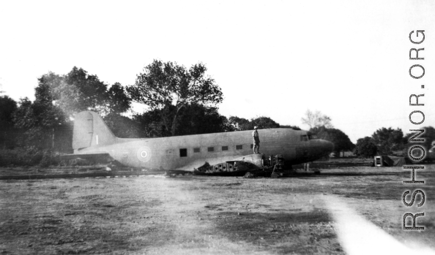A man in pith hat stands of fuselage remains of a C-47 transport next to railroad track. In the CBI during WWII.