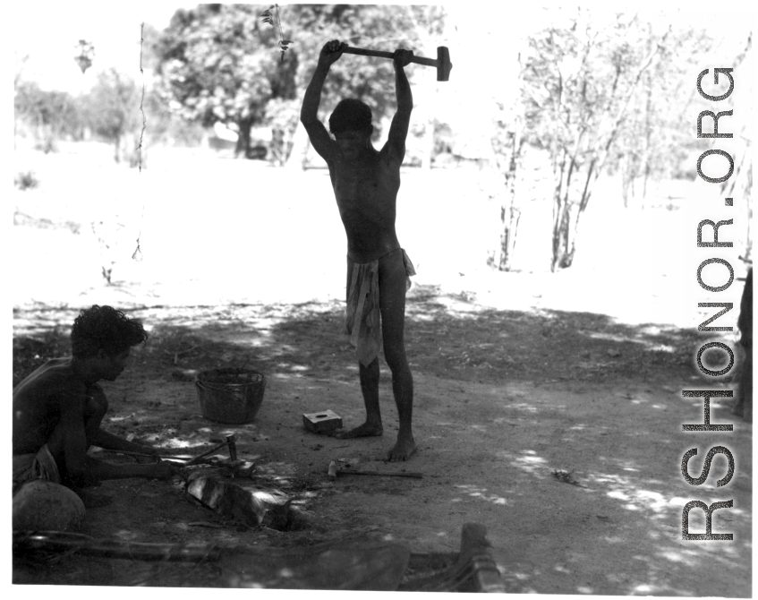 Men working metal in shade of tree.  Scenes in India witnessed by American GIs during WWII. For many Americans of that era, with their limited experience traveling, the everyday sights and sounds overseas were new, intriguing, and photo worthy.