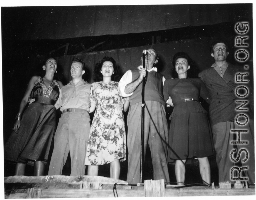Performers well off the beaten track, providing a break for Burma Road engineers of the 797th Engineer Forestry Company--Pat O'Brien and Jinx Falkenburg perform--Falkenburg, Yeaton, O'Brien, Carol, Dodd, and a random, confused-looking GI singing on stage.  During WWII.