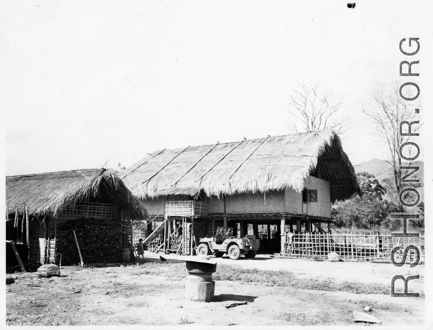 A child stands before a thatch house on stilts in Burma, with a jeep parked nearby.  Near the 797th Engineer Forestry Company.  During WWII.