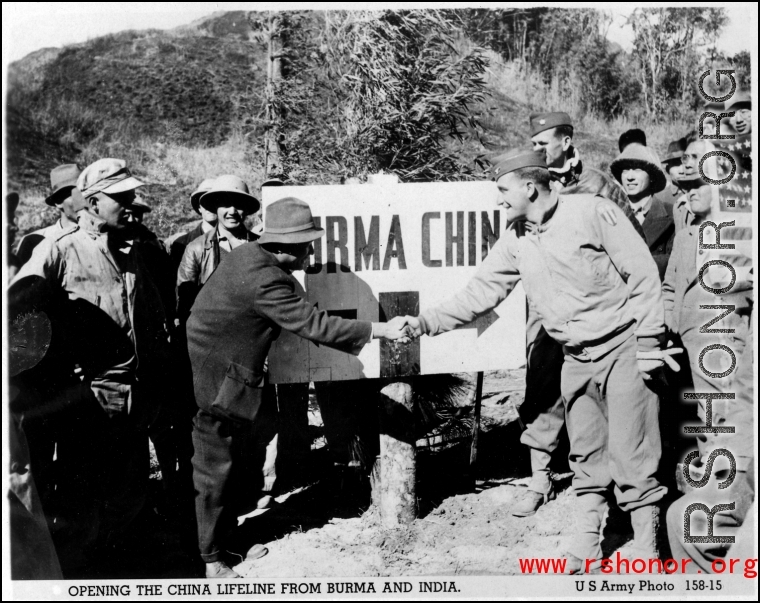 A GI and and a local man shake hands over the China-Burma border, a sign marking that boundary behind them, upon the opening of a road into China. During WWII.