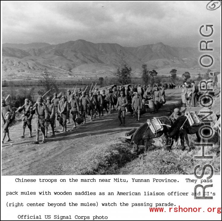 Chinese troops march near Mitu, Yunnan province, during WWII. They pass pack mules with wooden saddles as an American liaison officer and GIs (right center beyond mules) watch the passing parade.  US Signal Corps Photo.