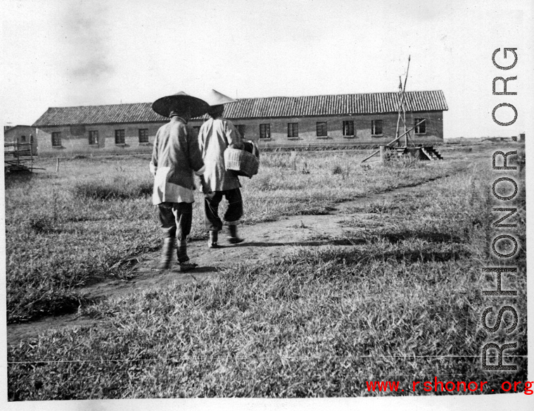 Two women with bound feet walking at an American base in Yunnan province, China, during WWII, with a barracks or other building in the background.