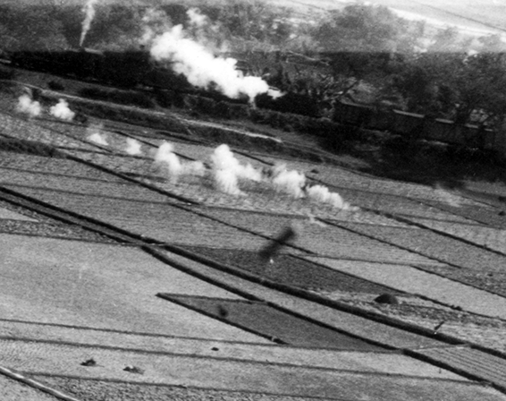 In the foreground, too close to be in focus, are a number of long cylinders in the air--presumably these are spent .50 caliber bullet cases falling from the B-25 as it shoots at the train.