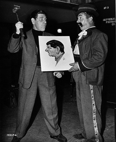Shemp Howard (left) visits Don Barclay, who holds a caricature, on the Universal Pictures set of Frankenstein Meets the Wolf Man (1943).