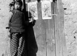 Local girl Shu Mei Chow stand before door decorated with door gods near the time of Chinese New Year in the village of Siang Shi Tsah, during WWII. Some of these were put out by the OWI in color.