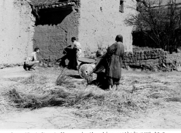 A GI points his carbine down the narrow space between two homes in a Chinese village during WWII, as a local village woman threshes grain.