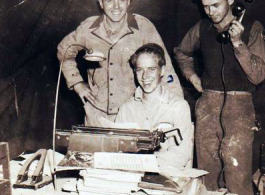 "Jim Kruger (standing), Elliot Knecht (seated), and an unknown member of the 396th having their picture taken in the Orderly Room."