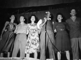 Performers well off the beaten track, providing a break for Burma Road engineers of the 797th Engineer Forestry Company--Pat O'Brien and Jinx Falkenburg perform--Falkenburg, Yeaton, O'Brien, Carol, Dodd, and a random, confused-looking GI singing on stage.  During WWII.