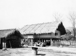 A child stands before a thatch house on stilts in Burma, with a jeep parked nearby.  Near the 797th Engineer Forestry Company.  During WWII.