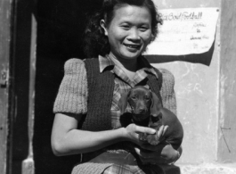 Alice Chong outside the HQ building at Kunming in 1944, holding the Dachshund puppy that her roommate Eloise Witwer gave to General Chennault.  Chennault named the dog “Little Joe” and then changed its name to “Joe-Dog” after it grew up.  The name, of course, was a way of getting back at his nemesis, General Joseph Stilwell because, as Chennault would often say, “Both were low-down, sons of bitches”. . .  Note the notice for the upcoming “Rice Bowl” football game pinned to the door frame too.  This is likel