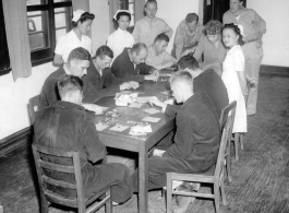 A crowd of servicemen in robes and in uniforms sit around a table playing bingo, with three Chinese nurses looking on in curiosity.