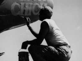 A GI paints the nickname "Slick Chick" on the nose of an aircraft, probably a photo reconnaissance P-38 (note the glass panel for a camera on the bottom of the nose).
