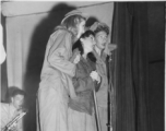 Ladies sing during USO performance in the CBI during WWII. GI plays sax by stage prop labeled for the 748th Railway Operating Battalion.