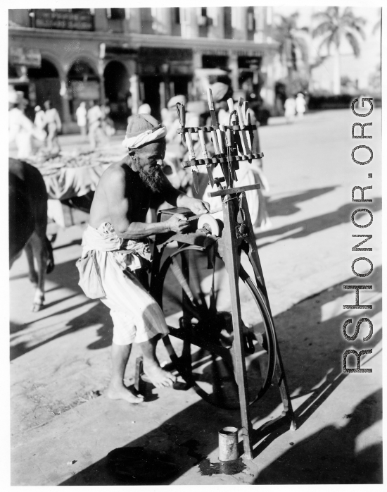 Man sharpens knives on road in town in Burma or India.  Near the 797th Engineer Forestry Company.  During WWII.