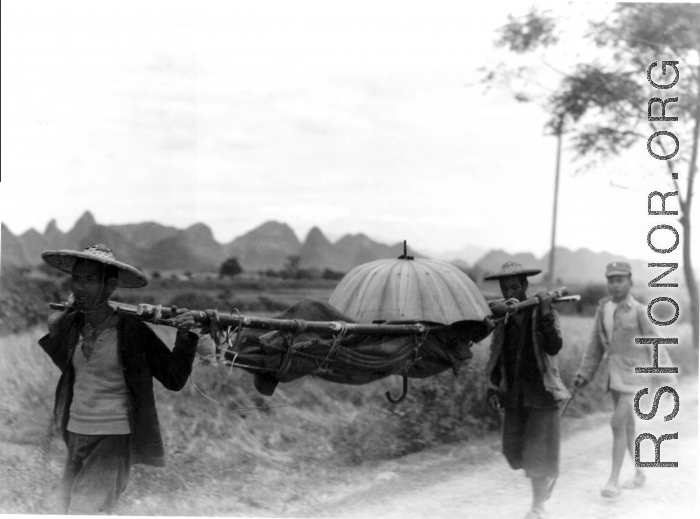 Men shoulder umbrella-covered stretcher with injured or sick person in Guangxi, China, during WWII.