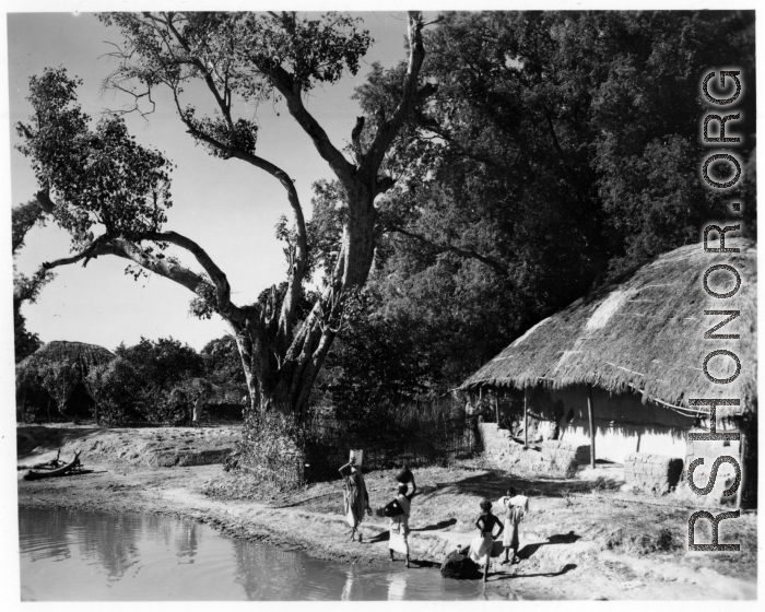 Women collect water from pond in front of house with thatched roof.  Scenes in India witnessed by American GIs during WWII. For many Americans of that era, with their limited experience traveling, the everyday sights and sounds overseas were new, intriguing, and photo worthy.