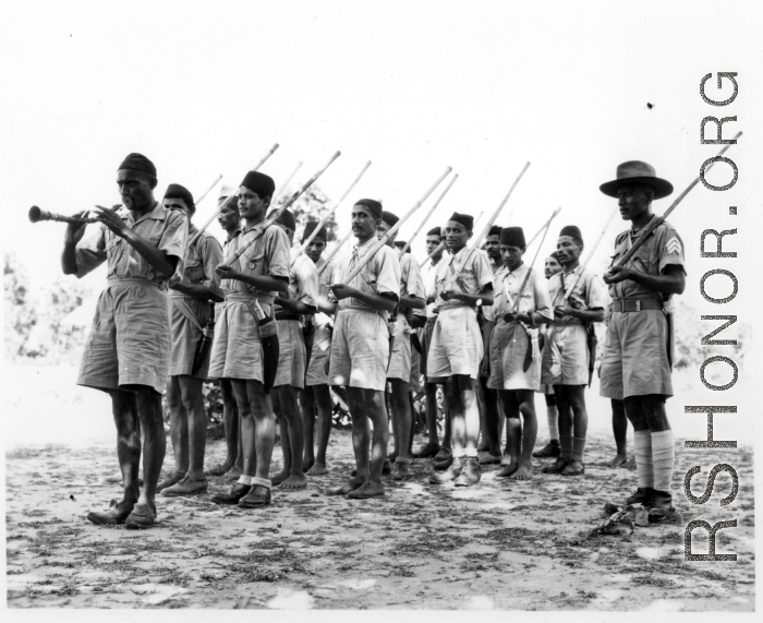 Indian soldiers march being led by a man blowing a horn, with wooden poles substituting for rifles.  Scenes in India witnessed by American GIs during WWII. For many Americans of that era, with their limited experience traveling, the everyday sights and sounds overseas were new, intriguing, and photo worthy.