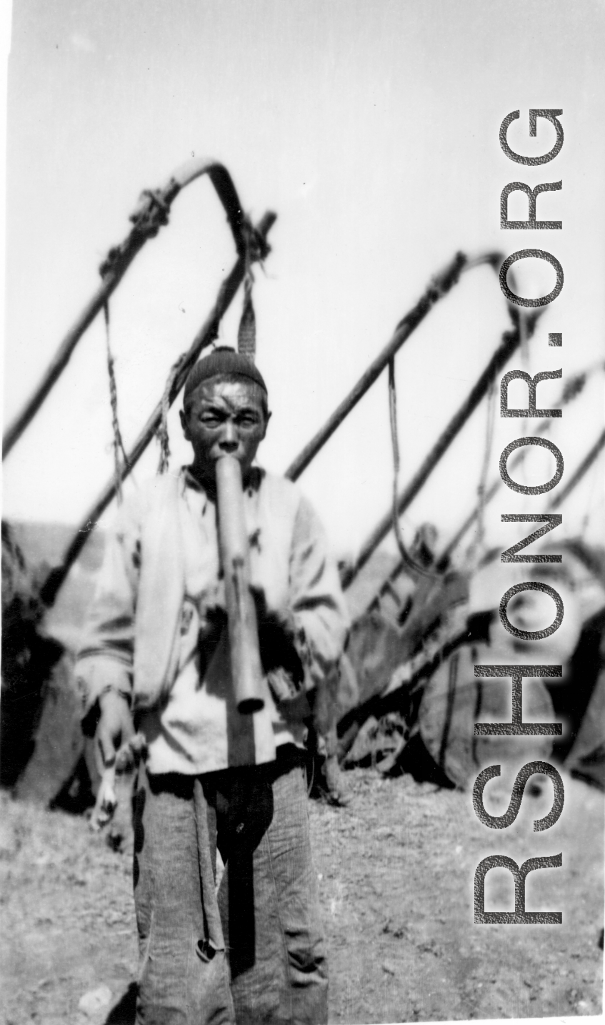 A Chinese man standing in front a row of donkey carts smokes from a water pipe. In China during WWII.