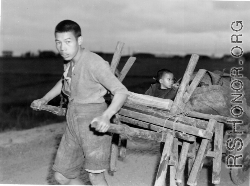 In the near darkness a man in China pulls a cart loaded with benches, an oven in a 55-gallon drum, a scale, and a son--a retail stand or eatery being taken out for a day of work, or being brought back after a day of work. During WWII.