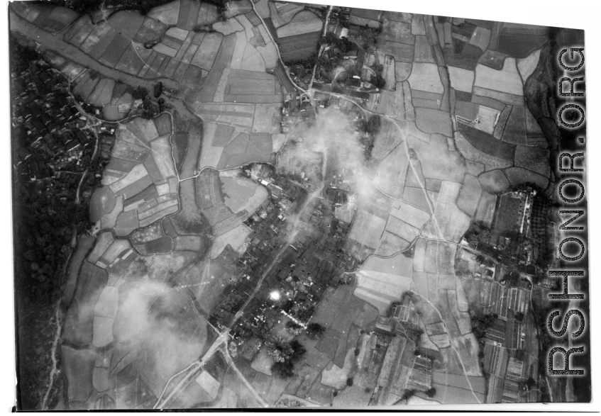 Bombs explode on a compact village in the midst of rice paddies in either SW China (esp. Guangxi), or Burma, or French Indochina during WWII. The bright white spot is the arc flash of a bomb explosion, caught exactly at the right time.