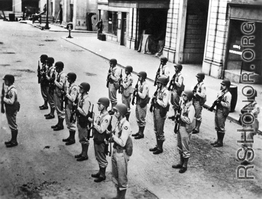 GIs, with apparent Army Air Forces shoulder patches, stand in ranks in a place that uses the word "haberdashery" (as on shop window to the right)--likely India, but also possibly Britain.