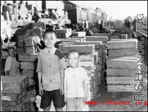 Two refugee children pose for the photographer before crates of supplies. At the train station in Liuzhou during WWII, in the fall of 1944, as the Japanese advanced during the Ichigo campaign.