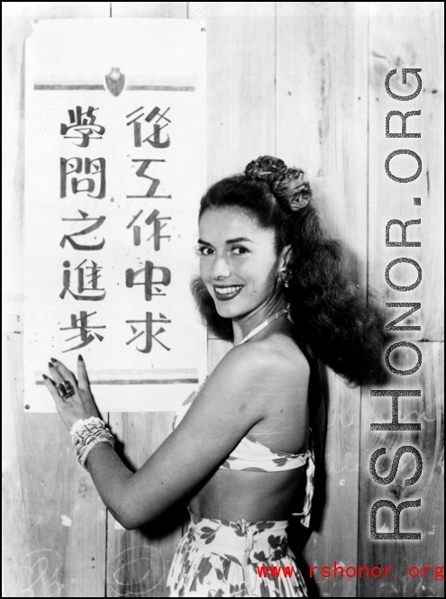 Jinx Falkenburg poses with a classroom slogan that says, roughly, "from cooperation seek progress in learning." In China during WWII.