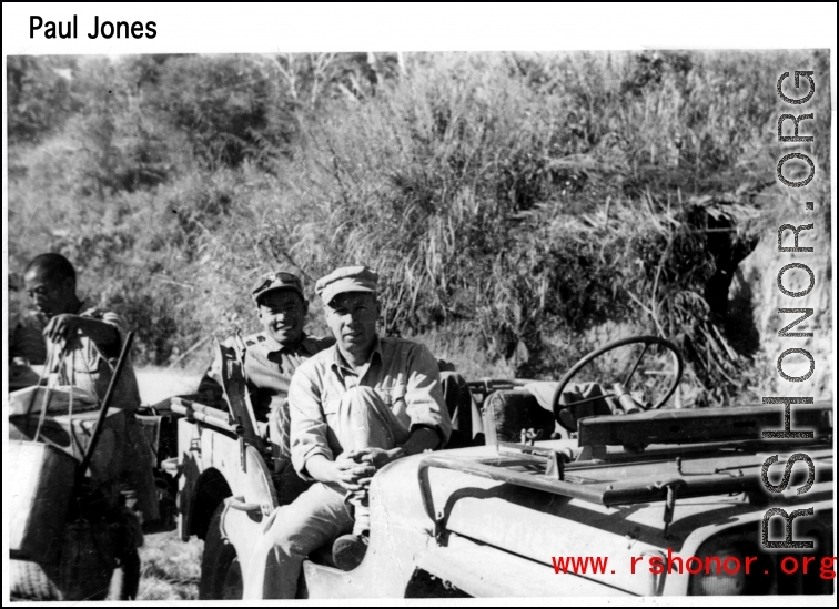 GI and Chinese soldier sit in a jeep, as a worker carries gear during WWII. Probably in Yunnan, China.