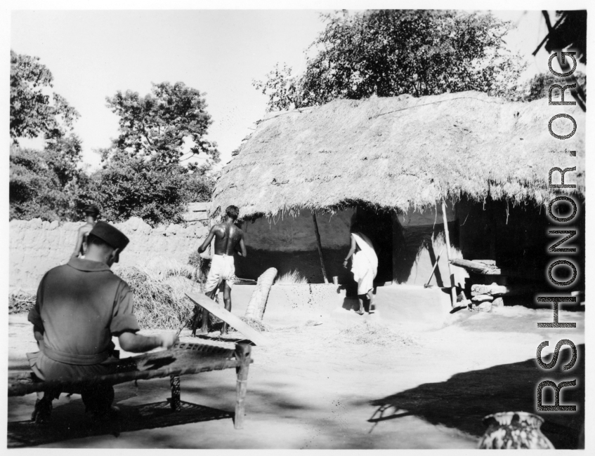 Soldier sitting on cot while local people go about chores.  Scenes in India witnessed by American GIs during WWII. For many Americans of that era, with their limited experience traveling, the everyday sights and sounds overseas were new, intriguing, and photo worthy.