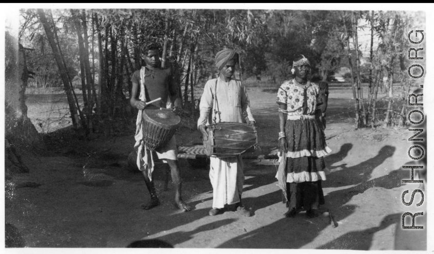 Men with cymbal, drum, and person in a dress.  Scenes in India witnessed by American GIs during WWII. For many Americans of that era, with their limited experience traveling, the everyday sights and sounds overseas were new, intriguing, and photo worthy.