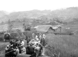 Chinese men eat a banquet, while GIS stand to one side and look at something. The white bands on the heads of some of the men who are eating implies this is a funeral banquet. In China during WWII.