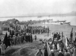 A large crowd along a river in China during WWII, gathered for some kind of communal activity. On the right is a dragon dance team, and numerous people hold banners and flags.