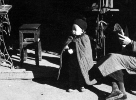 A young boy stands in the doorway of a seamstress shop in China, dressed in a warm cape. During WWII.