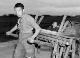 In the near darkness a man in China pulls a cart loaded with benches, an oven in a 55-gallon drum, a scale, and a son--a retail stand or eatery being taken out for a day of work, or being brought back after a day of work. During WWII.