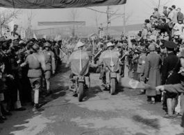 Burma Road dedication parade and ceremony in Kunming, China, on or around February 4, 1945, during WWII. Review of first convoy (or one of the first convoys) to reach China. Ranks of soldiers and civilians, waiting in anticipation of the parade, speeches, and first trucks to arrive, watch motorcycle MPs and jeeps, with US and Chinese flags.