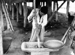 Local people in Burma near the 797th Engineer Forestry Company--A man is working most likely to dehusk grain, or to make some other worked produce, while another person waits with a woven dry pan.  During WWII.