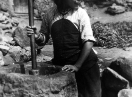A young woman uses a wooden rod to husk rice in a large stone mortar. Local people in China, probably in Yunnan province.   From the collection of Wozniak, combat photographer for the 491st Bomb Squadron, in the CBI.