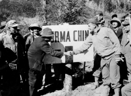 A GI and and a local man shake hands over the China-Burma border, a sign marking that boundary behind them, upon the opening of a road into China. During WWII.