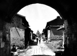 Looking into an old Chinese village through a gate, with a drum tower or bell tower in the distance, in the middle of the village. During WWII.