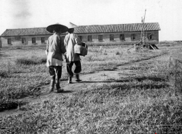 Two women with bound feet walking at an American base in Yunnan province, China, during WWII, with a barracks or other building in the background.