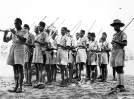 Indian soldiers march being led by a man blowing a horn, with wooden poles substituting for rifles.  Scenes in India witnessed by American GIs during WWII. For many Americans of that era, with their limited experience traveling, the everyday sights and sounds overseas were new, intriguing, and photo worthy.