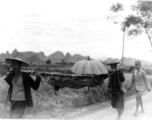 Men shoulder umbrella-covered stretcher with injured or sick person in Guangxi, China, during WWII.